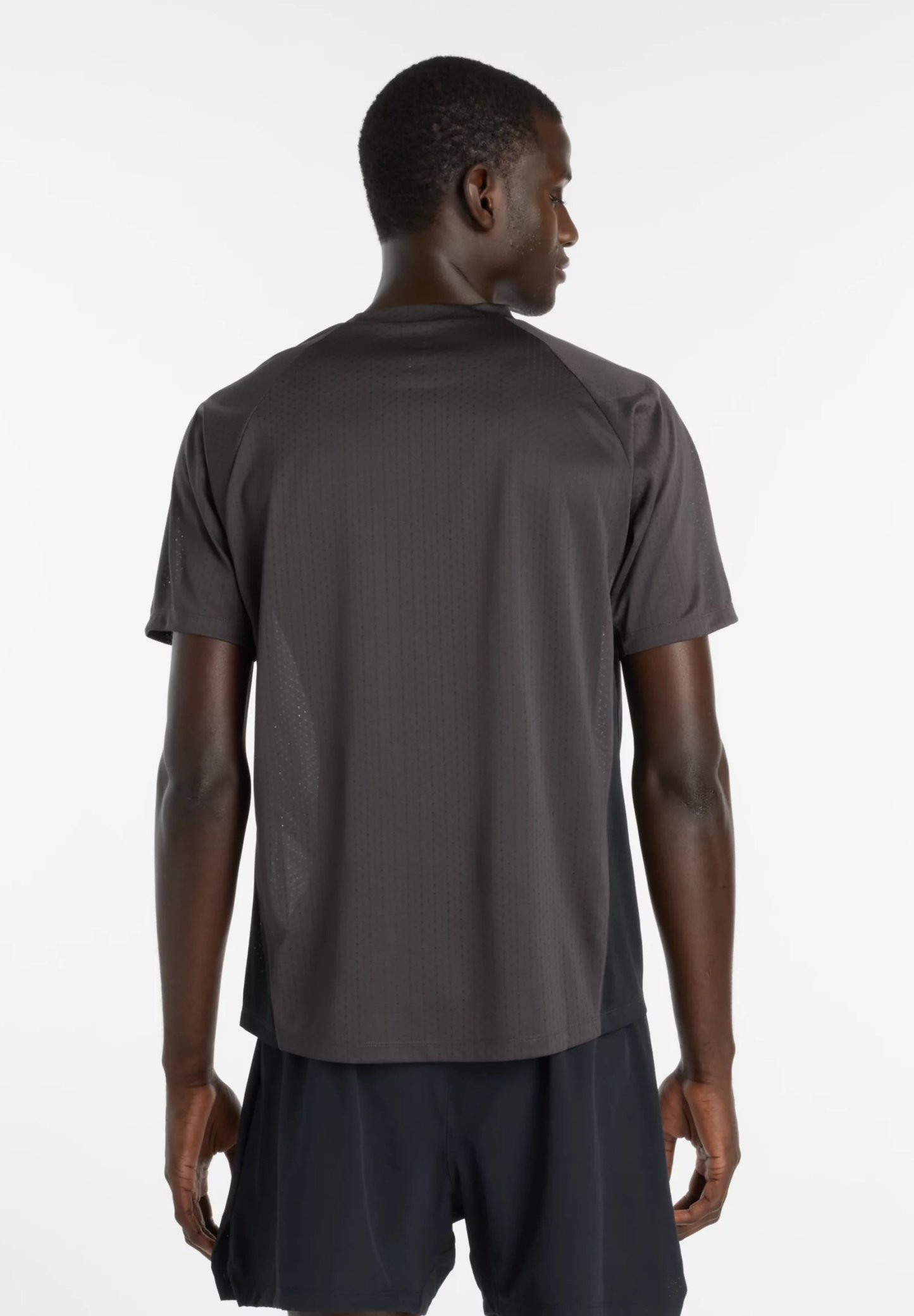 Hombre que lleva una camiseta de manga corta gris oscuro y pantalones cortos negros, de pie de espaldas sobre un fondo blanco.