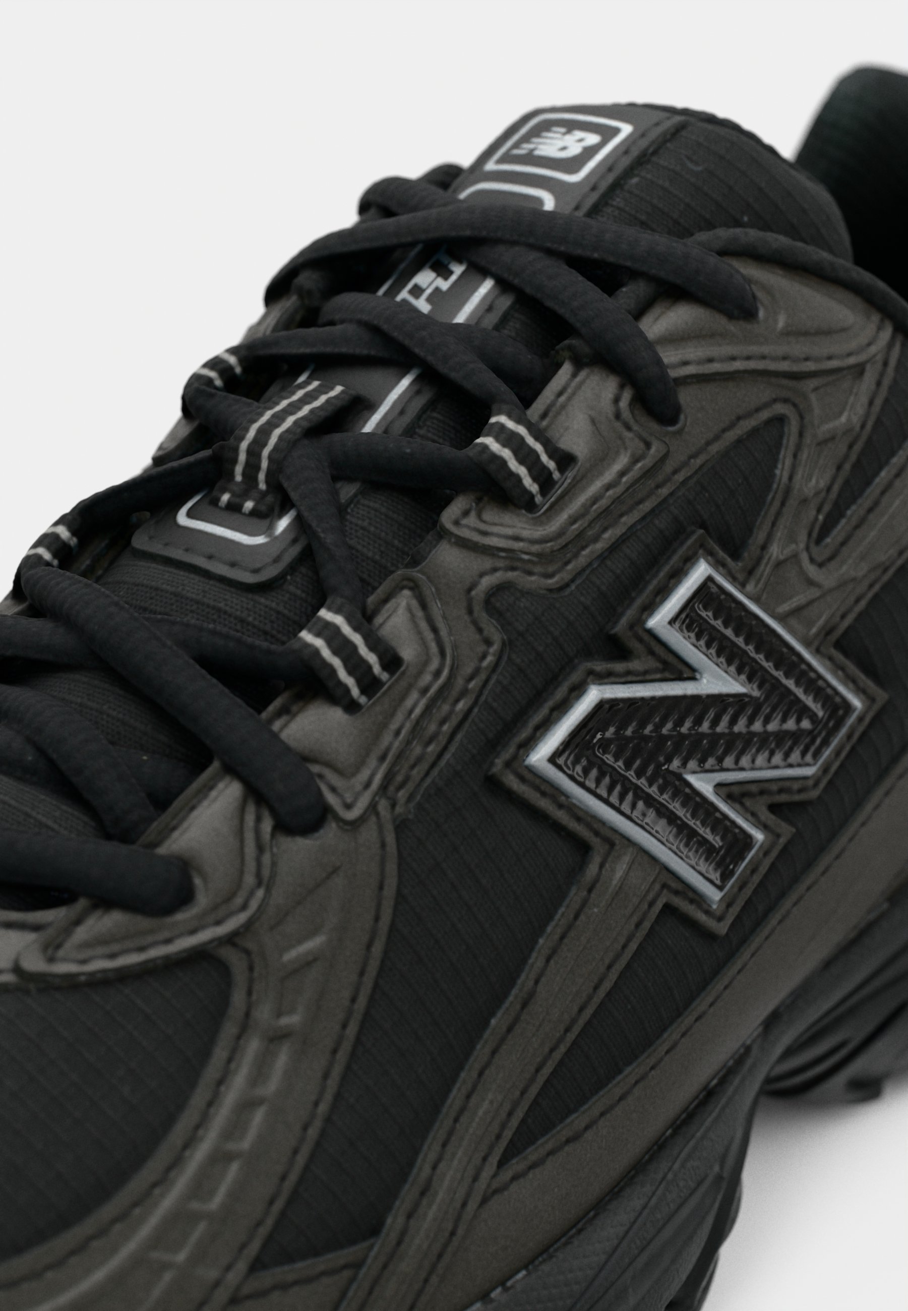 Zapatillas negras con una parte superior texturizada y suaves acanaladuras, un prominente logo 'N' plateado, y cordones planos negros con acentos blancos.
