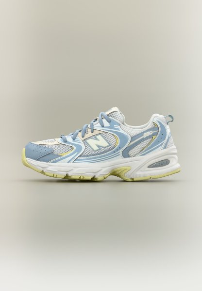 Zapatilla deportiva blanca y azul claro con tela de malla, cordones y detalles amarillos, mostrada de lado sobre un fondo neutro.