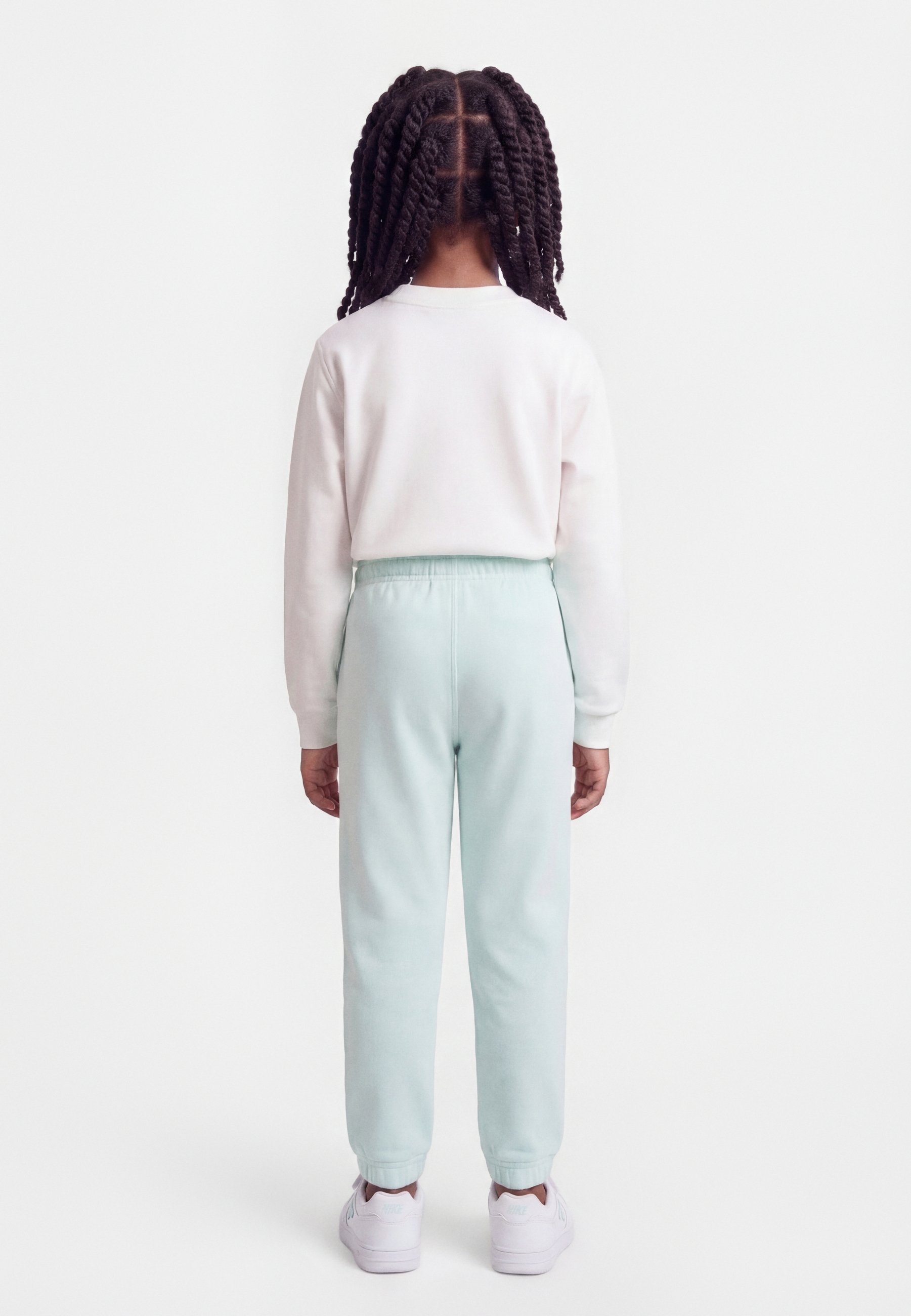 Niño con trenzas torcidas de espaldas, vistiendo una camisa blanca de manga larga, pantalones azul claro y zapatillas blancas sobre fondo blanco.