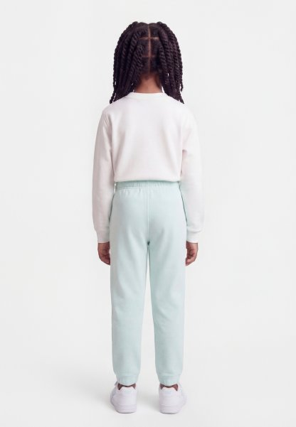 Niño con trenzas torcidas de espaldas, vistiendo una camisa blanca de manga larga, pantalones azul claro y zapatillas blancas sobre fondo blanco.