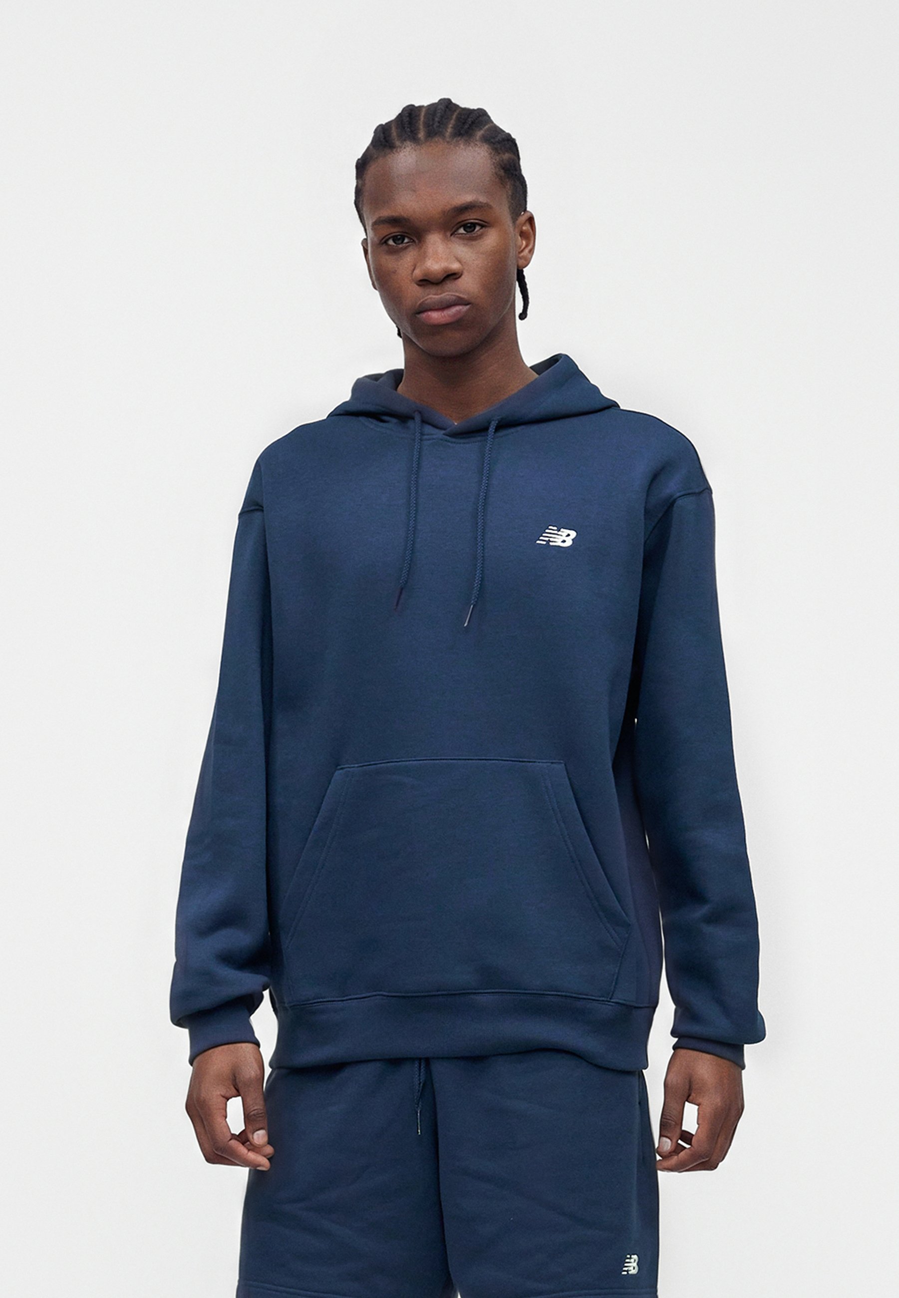 Persona joven con cabello trenzado que lleva una sudadera New Balance azul marino y pantalones cortos a juego, de pie sobre un fondo blanco liso.