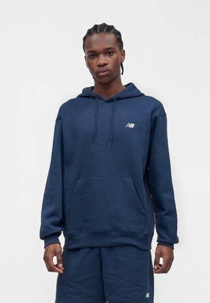 Persona joven con cabello trenzado que lleva una sudadera New Balance azul marino y pantalones cortos a juego, de pie sobre un fondo blanco liso.