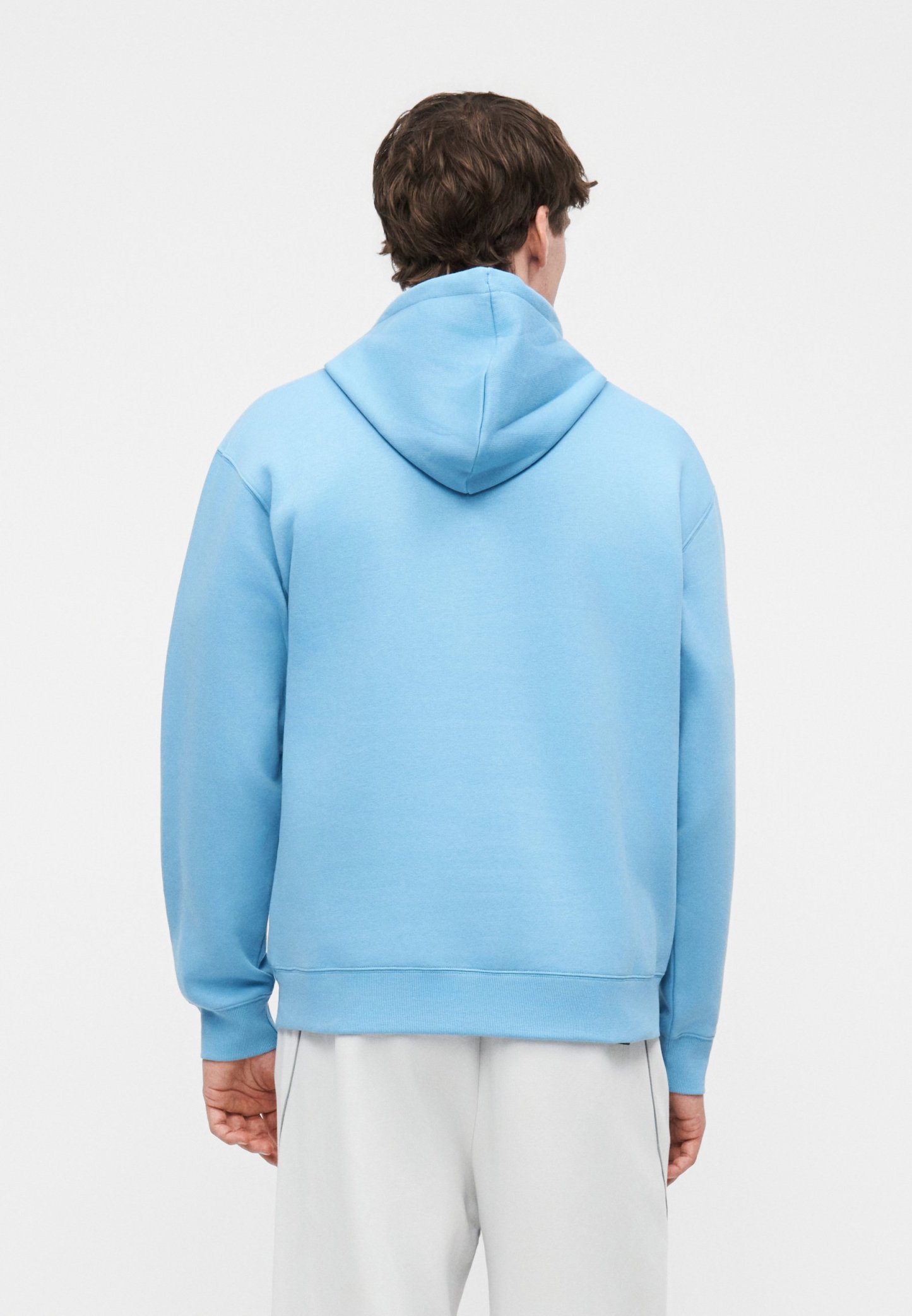 Hombre vestido con sudadera con capucha azul claro y pantalones blancos, de pie de espaldas sobre un fondo liso.