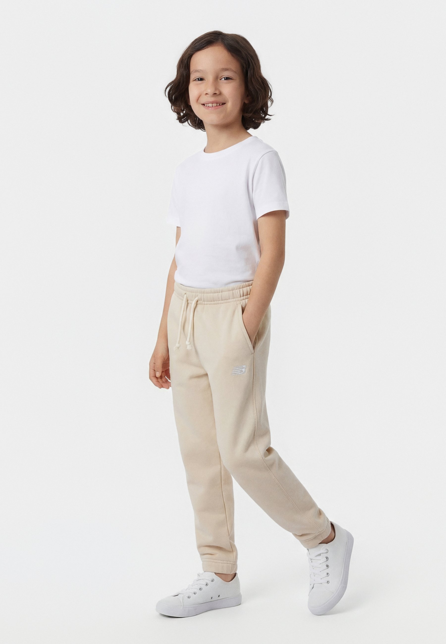 Niño sonriente con cabello rizado, vestido con una camiseta blanca, pantalones deportivos beige con cordón y zapatillas blancas, de pie con una mano en el bolsillo.