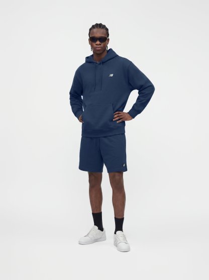 Hombre con sudadera con capucha azul marino y pantalones cortos, calcetines negros, zapatillas blancas y gafas de sol oscuras, de pie con las manos en las caderas.