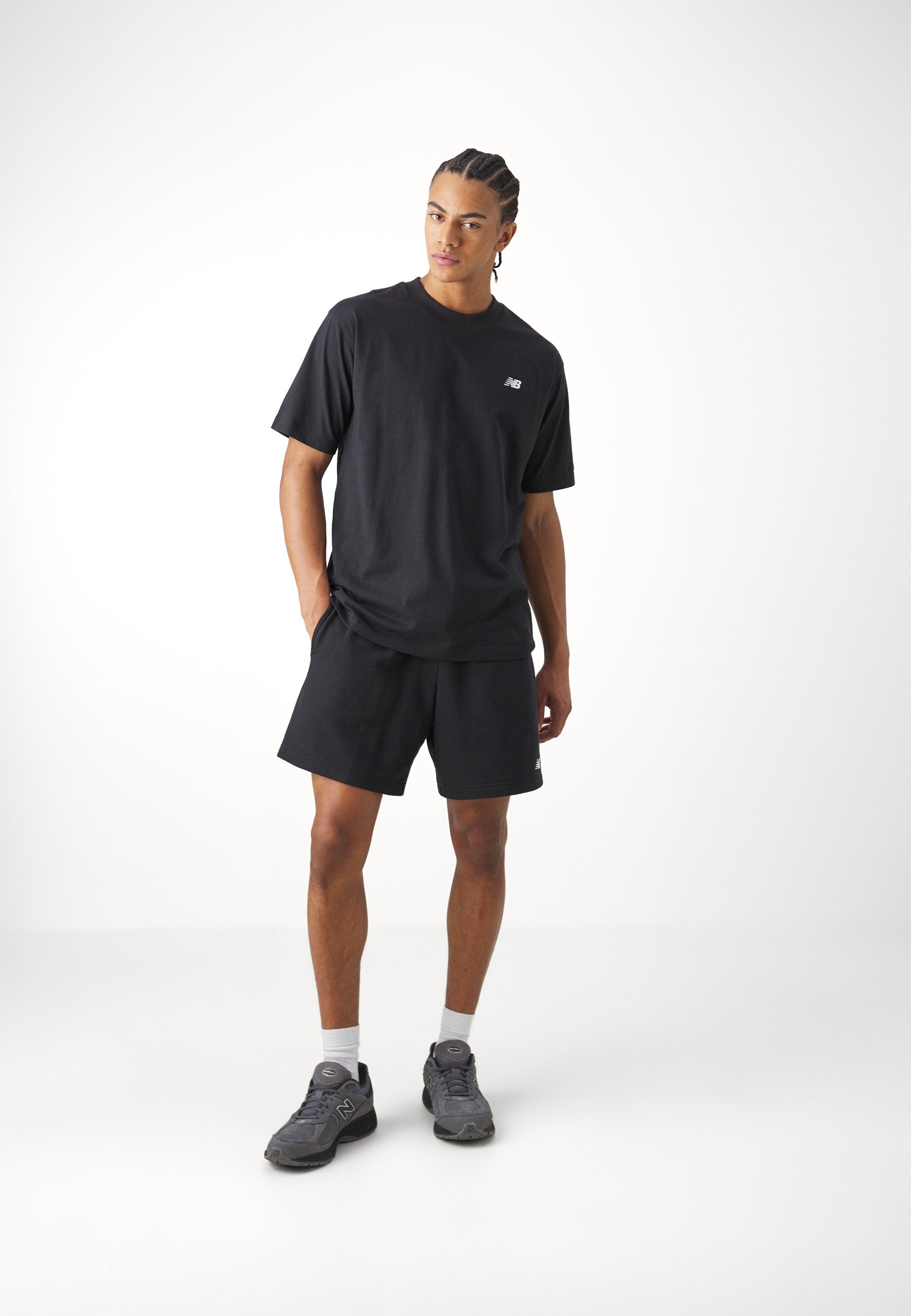 Camiseta de algodón negra con mangas cortas y un pequeño logo, combinada con pantalones cortos de algodón negros. Lleva zapatillas grises y calcetines blancos.
