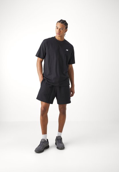 Camiseta de algodón negra con mangas cortas y un pequeño logo, combinada con pantalones cortos de algodón negros. Lleva zapatillas grises y calcetines blancos.