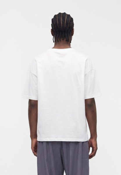 Persona con cabello trenzado lleva una camiseta blanca lisa de manga corta y pantalones grises, de pie de espaldas contra un fondo blanco.