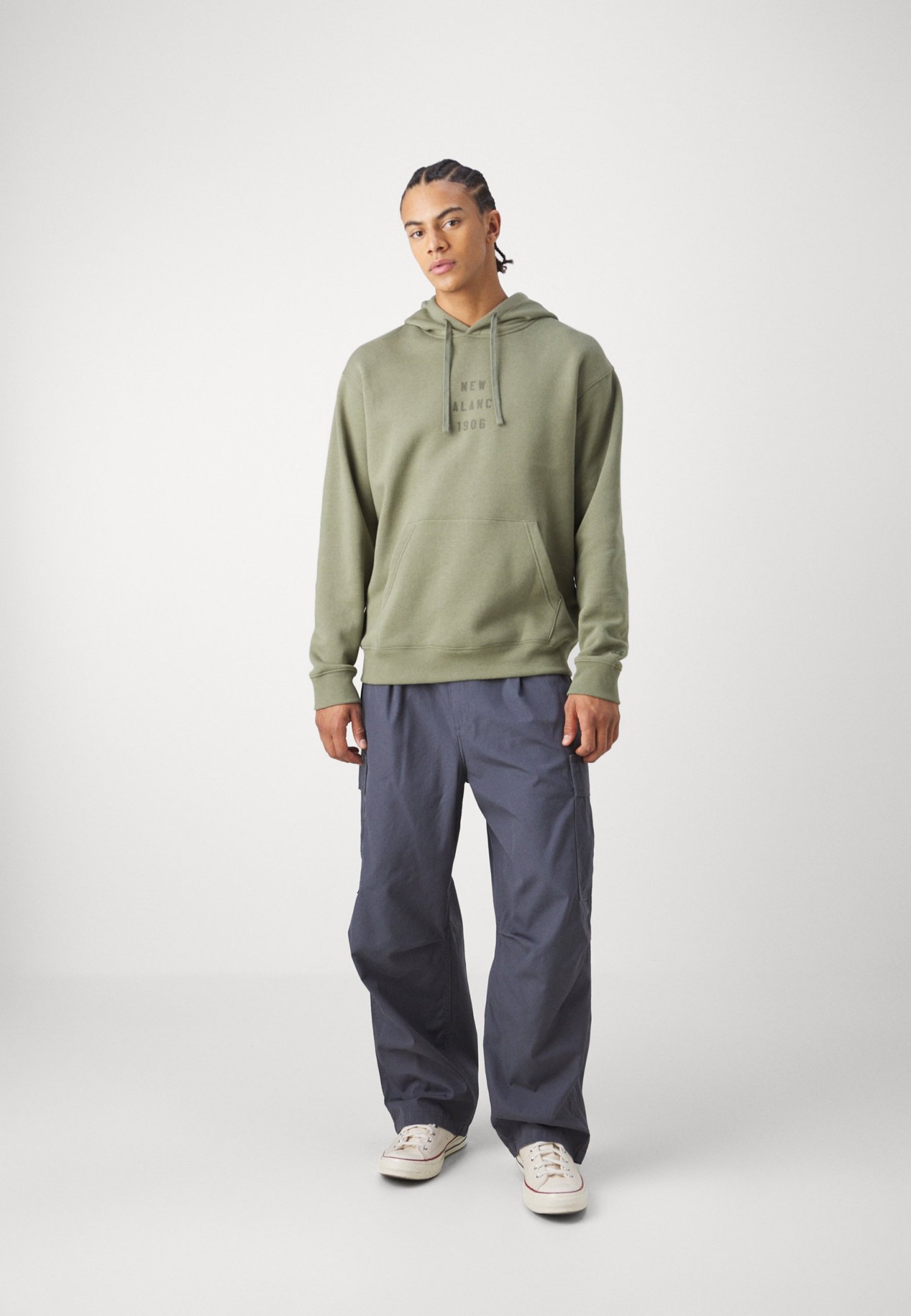 Sudadera con capucha de color verde oliva con bolsillo frontal y logo, combinada con pantalones cargo de corte holgado en gris oscuro y zapatillas blancas.