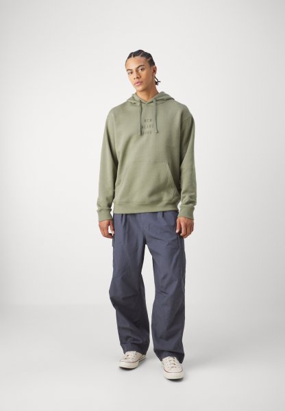 Sudadera con capucha de color verde oliva con bolsillo frontal y logo, combinada con pantalones cargo de corte holgado en gris oscuro y zapatillas blancas.