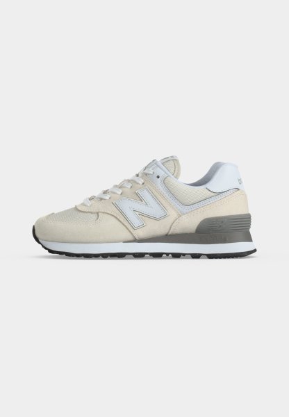 Zapatilla New Balance beige y blanca con parte superior de malla y ante, soporte de talón gris y suela de goma negra, mostrada de perfil.