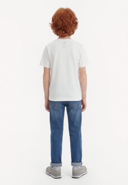 Niño con cabello rizado y rojo, vestido con una camiseta blanca lisa, vaqueros azules y zapatillas gris New Balance, de pie de espaldas sobre fondo blanco.