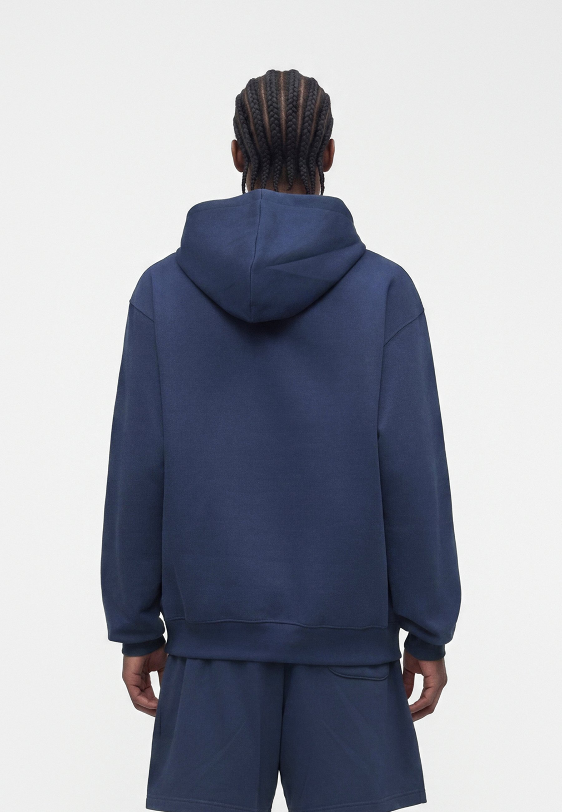 Persona con cabello trenzado que lleva una sudadera azul marino con capucha y pantalones cortos a juego, de espaldas sobre un fondo blanco liso.