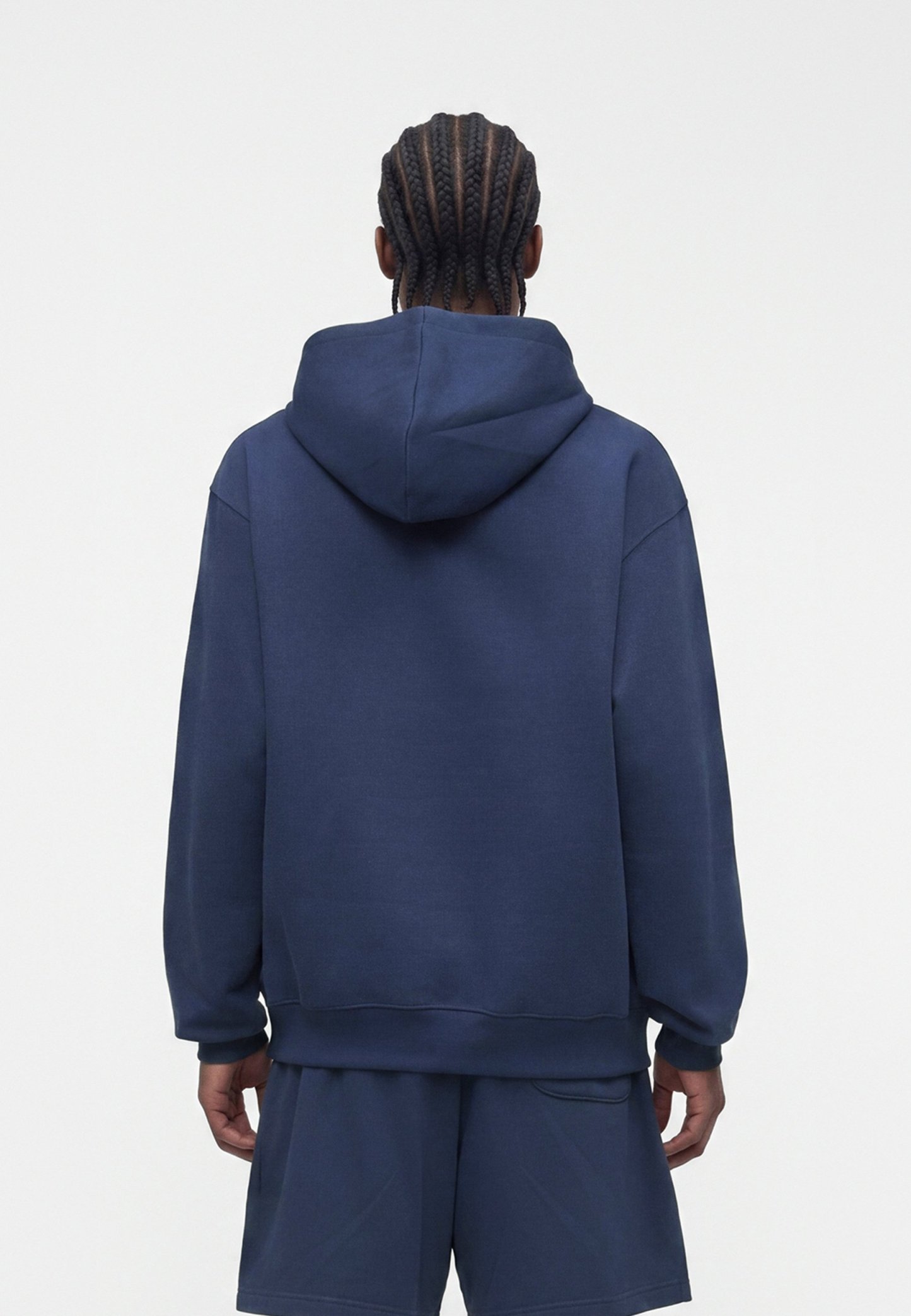 Persona con cabello trenzado que lleva una sudadera azul marino con capucha y pantalones cortos a juego, de espaldas sobre un fondo blanco liso.