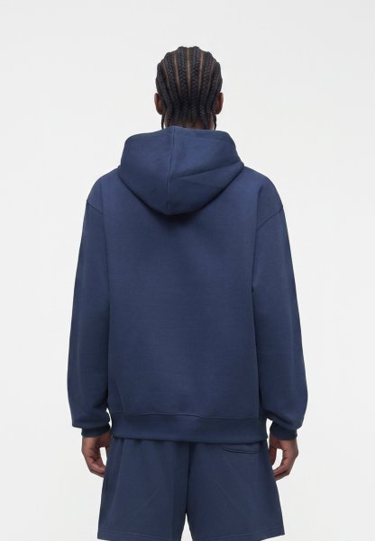 Persona con cabello trenzado que lleva una sudadera azul marino con capucha y pantalones cortos a juego, de espaldas sobre un fondo blanco liso.