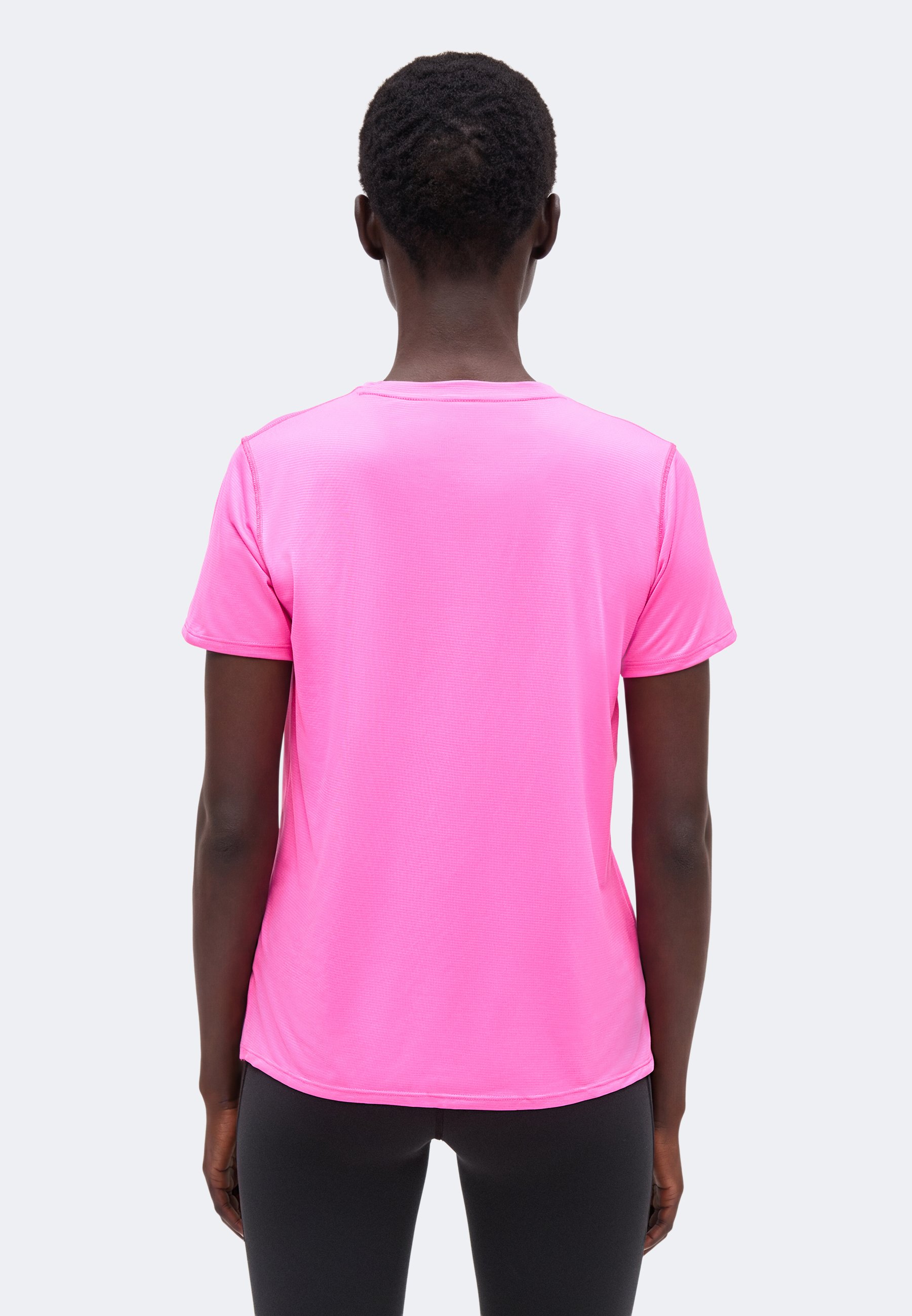 Persona con cabello corto que lleva una camiseta de manga corta de color rosa brillante y pantalones negros, de pie de espaldas sobre un fondo liso.