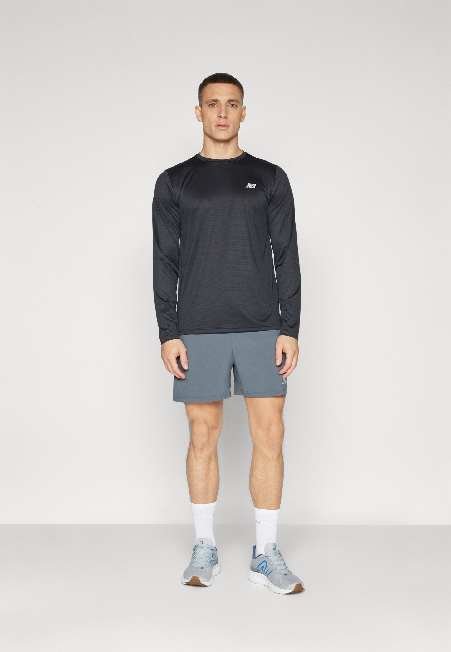 Conjunto deportivo para hombre que incluye una camiseta negra de manga larga y pantalones cortos grises. Zapatillas grises con detalles azules, calcetines blancos y un diseño simple.