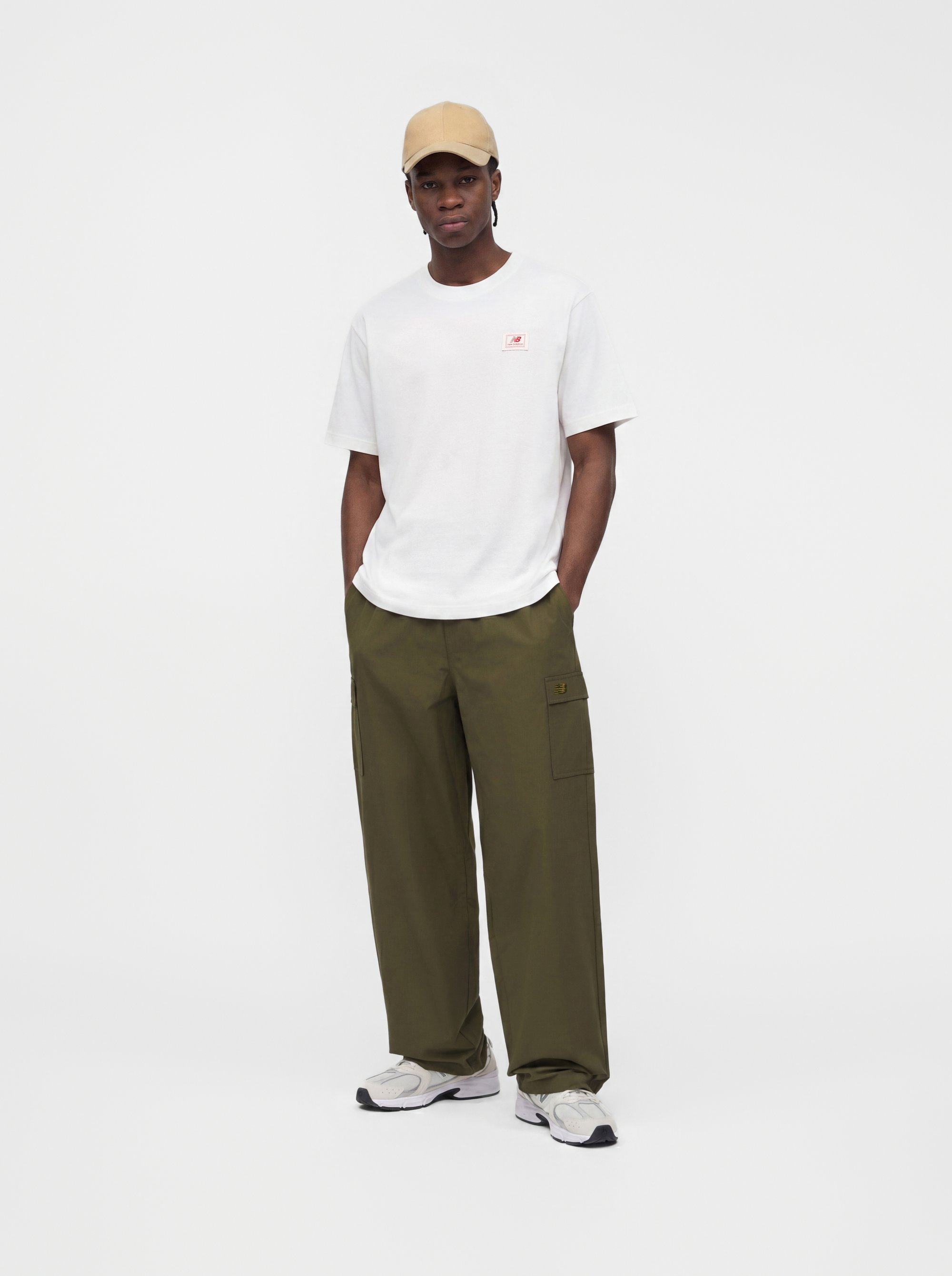 Hombre con gorra beige, camiseta blanca de manga corta, pantalones cargo verde oliva y zapatillas blancas, de pie con las manos en los bolsillos.