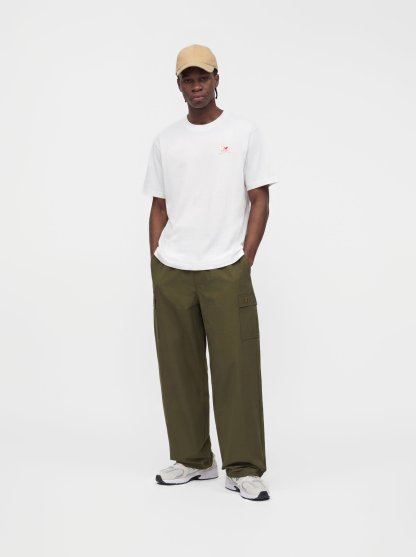Hombre con gorra beige, camiseta blanca de manga corta, pantalones cargo verde oliva y zapatillas blancas, de pie con las manos en los bolsillos.