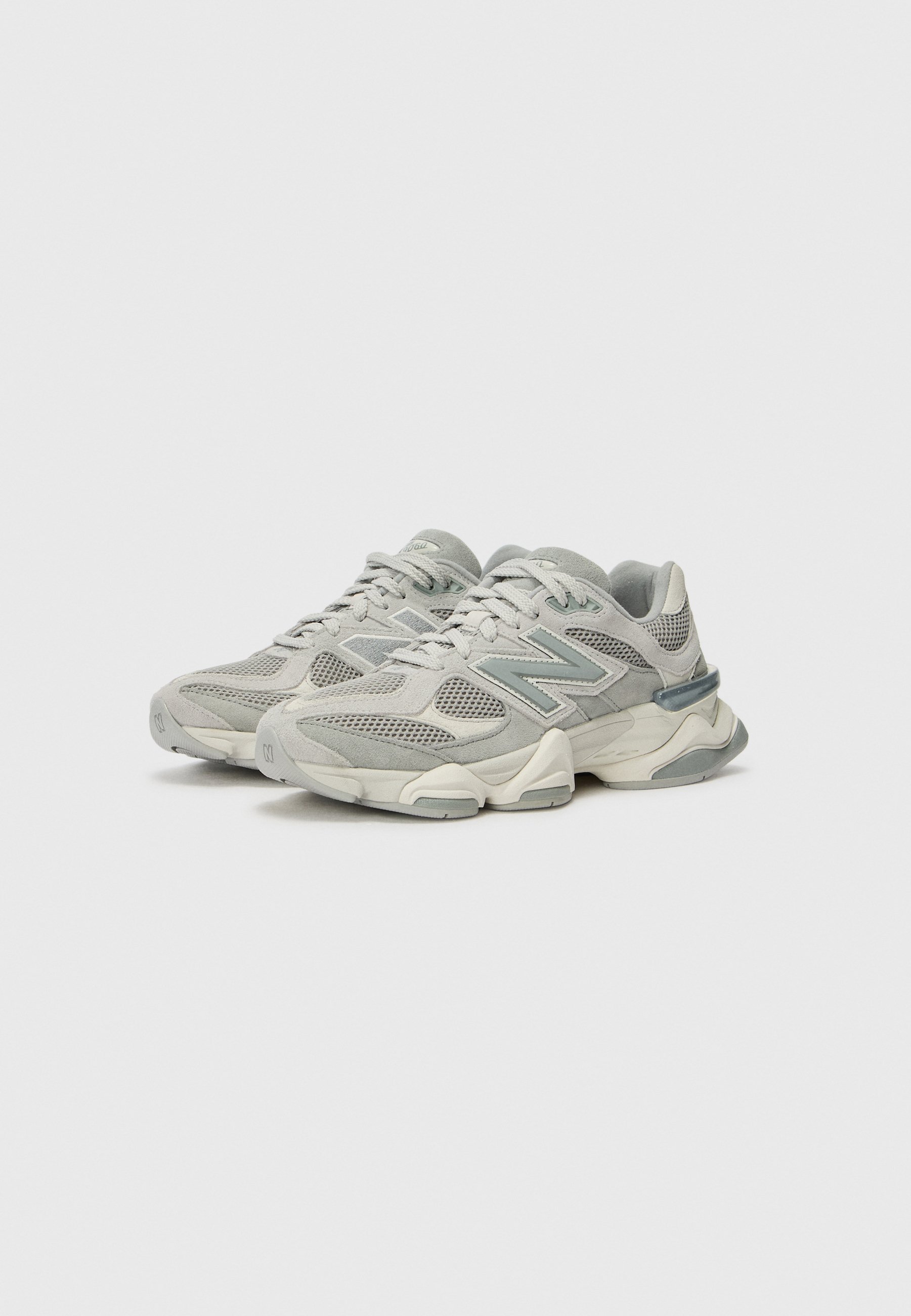 Par de zapatillas New Balance en gris y blanco roto con paneles de malla y ante, suela gruesa y cierre de cordones, mostradas una al lado de la otra.