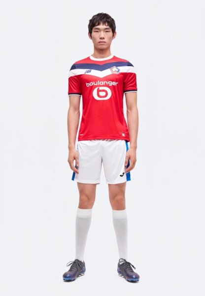 Un joven está de pie vistiendo una camiseta roja del Lille, pantalones cortos blancos, calcetas blancas hasta la rodilla y botas negras sobre un fondo blanco liso.