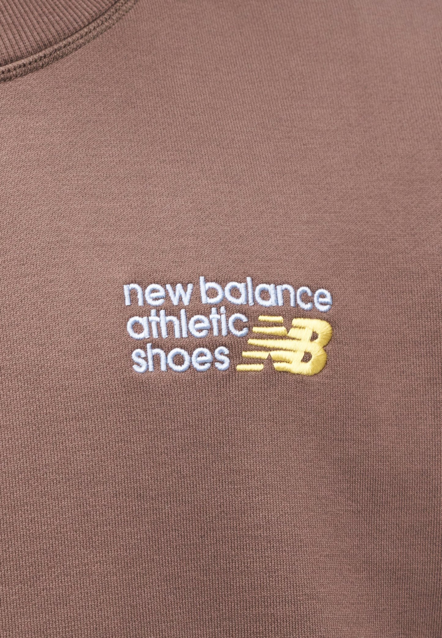 Textil marrón con la inscripción "new balance athletic shoes" bordada en blanco con un acento amarillo en un escote redondeado.