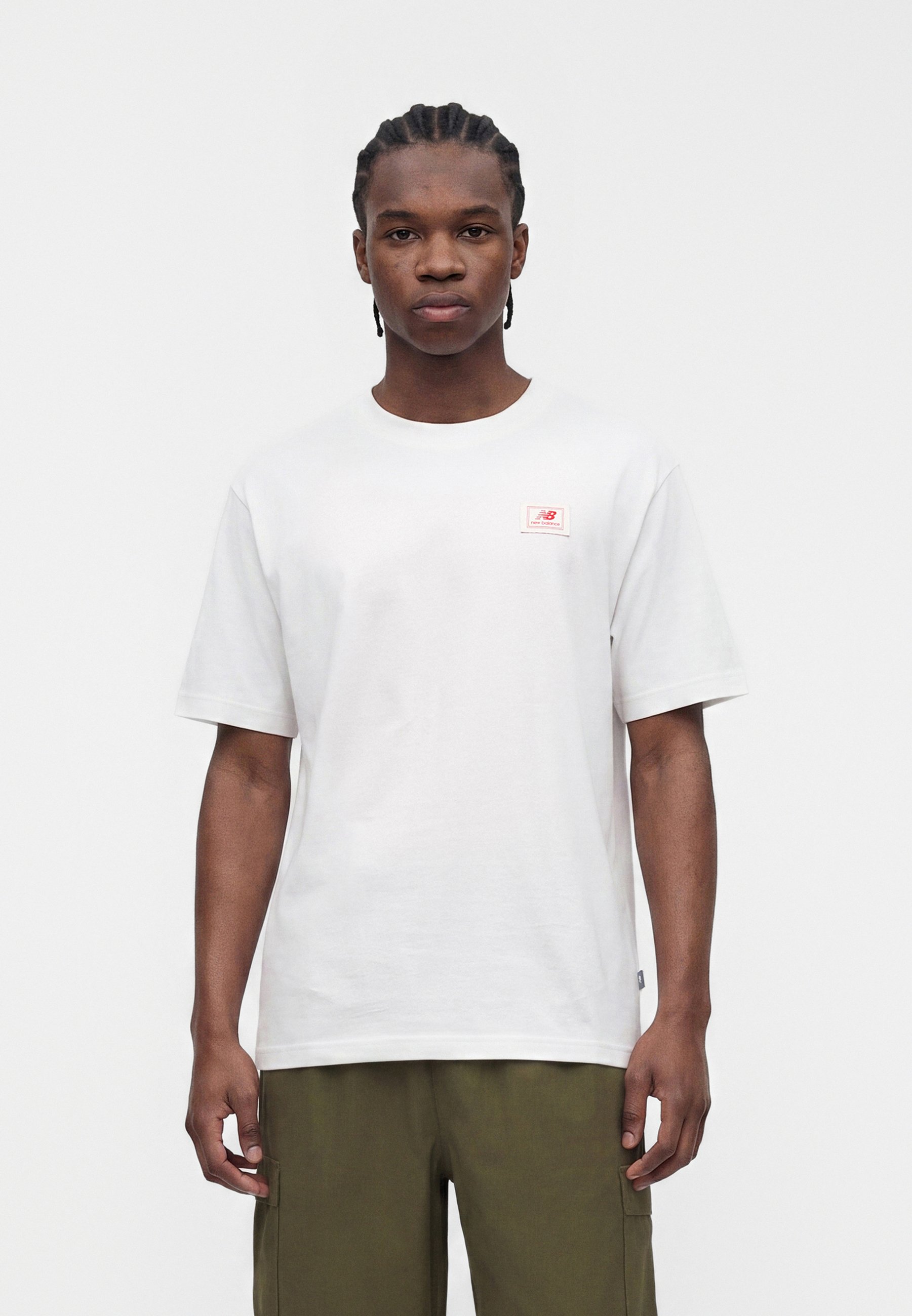 Joven con el pelo trenzado, con una camiseta blanca lisa y pantalones verde oliva, de pie frente a un fondo claro y liso.