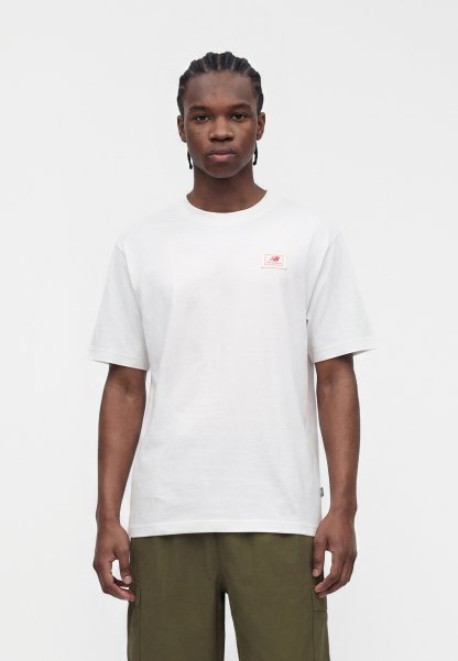 Joven con el pelo trenzado, con una camiseta blanca lisa y pantalones verde oliva, de pie frente a un fondo claro y liso.