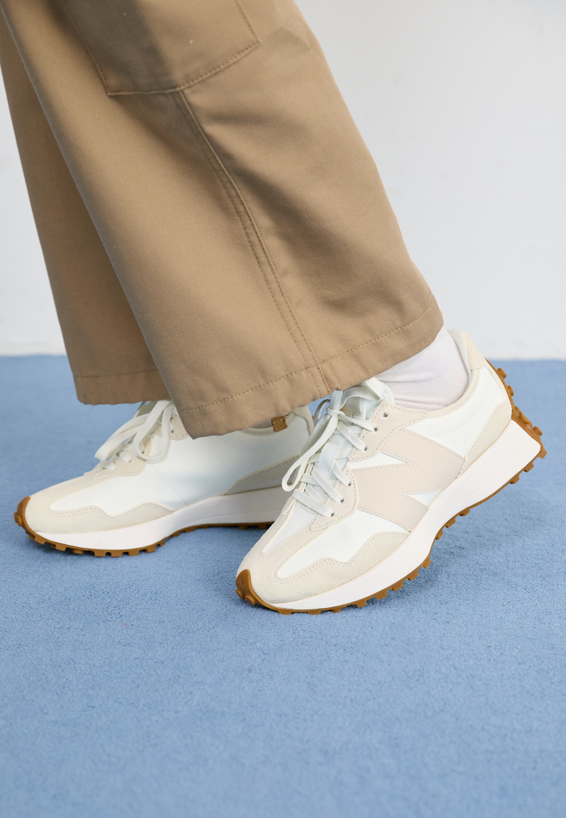 Zapatillas deportivas blancas y beige con superficies texturizadas, suela de goma y cordones azul claro, combinadas con pantalones beige de pierna ancha.