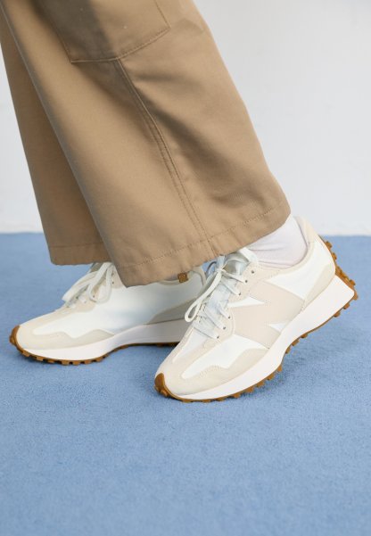 Zapatillas deportivas blancas y beige con superficies texturizadas, suela de goma y cordones azul claro, combinadas con pantalones beige de pierna ancha.