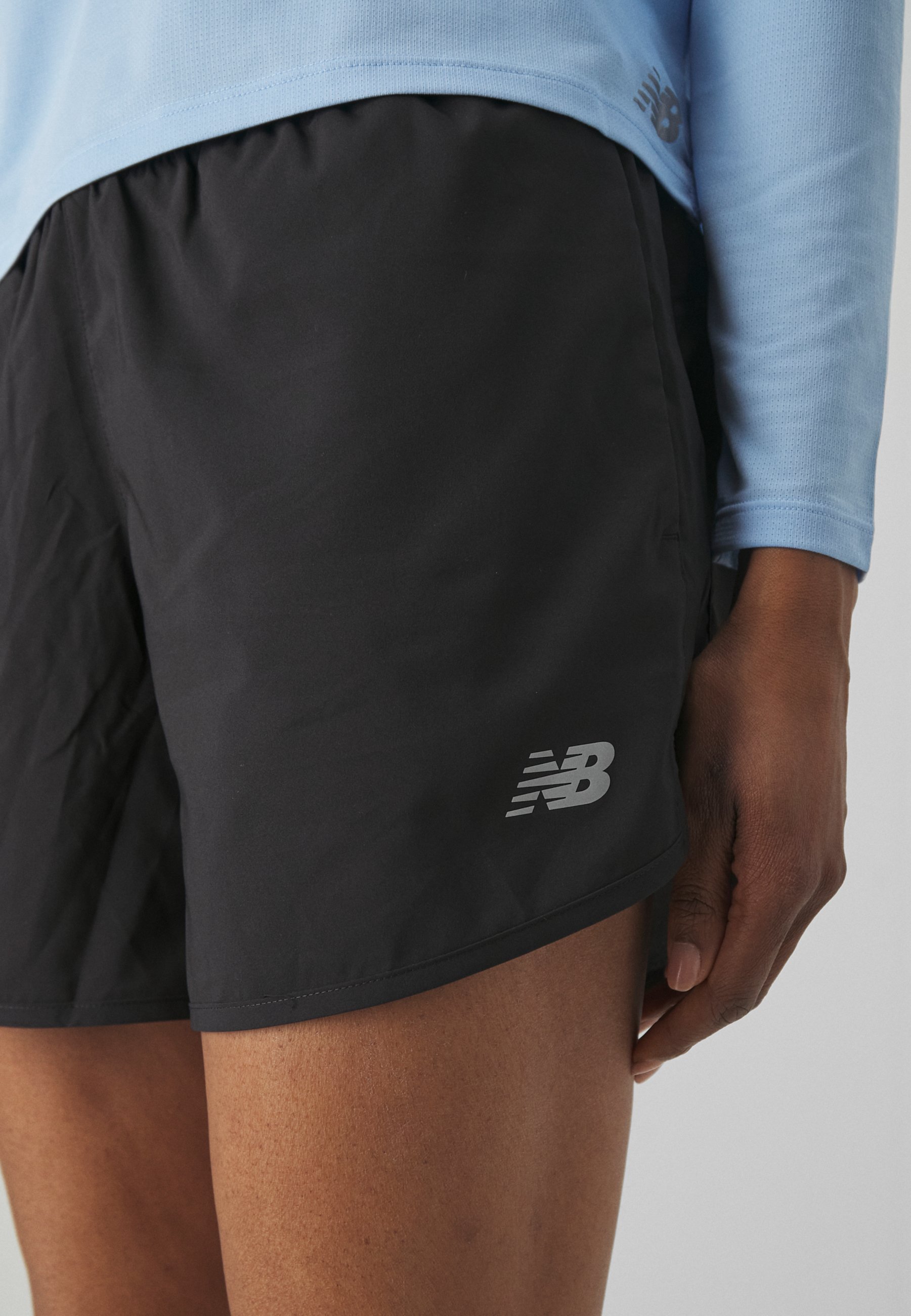 Pantalones cortos deportivos negros con una textura ligera y suave, que presentan un logo reflectante de New Balance en la parte inferior izquierda.