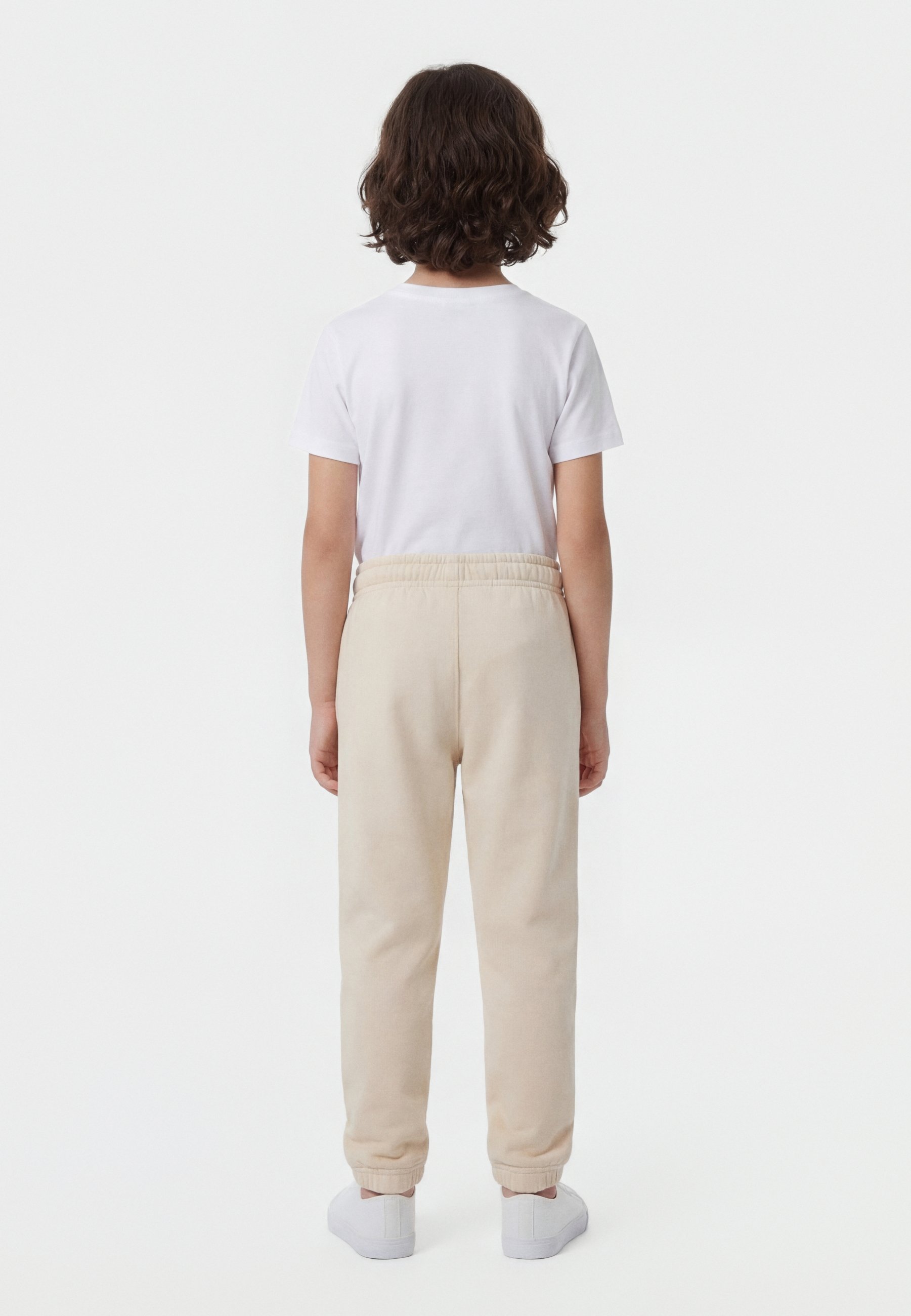 Niño con cabello rizado castaño, vestido con una camiseta blanca, pantalones deportivos beige y zapatillas blancas, de pie de espaldas sobre un fondo blanco.