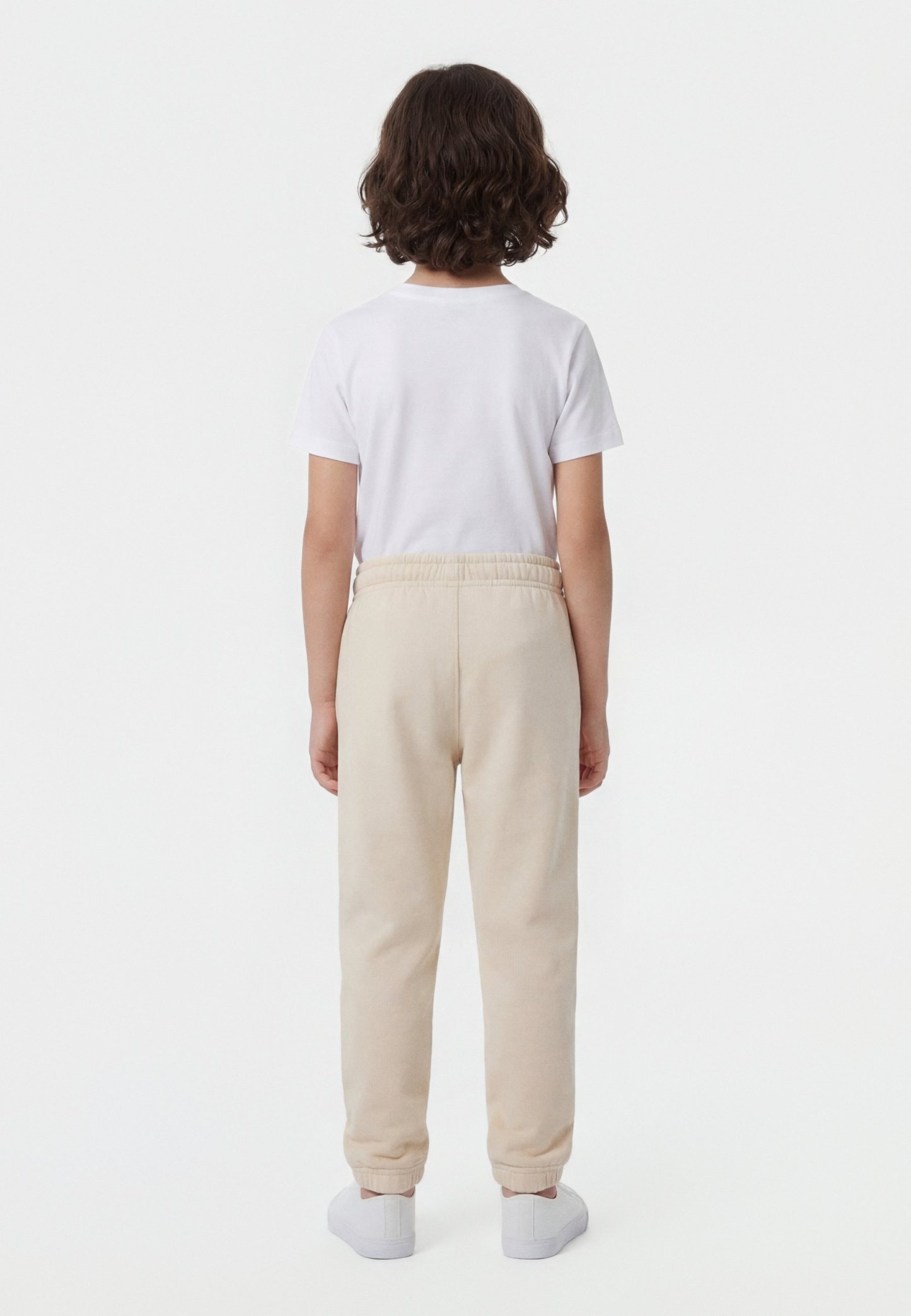 Niño con cabello rizado castaño, vestido con una camiseta blanca, pantalones deportivos beige y zapatillas blancas, de pie de espaldas sobre un fondo blanco.