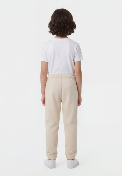 Niño con cabello rizado castaño, vestido con una camiseta blanca, pantalones deportivos beige y zapatillas blancas, de pie de espaldas sobre un fondo blanco.