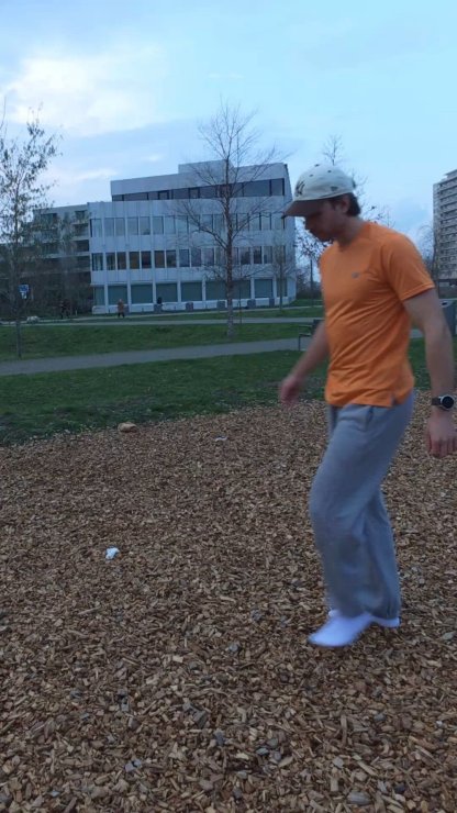 Hombre con camiseta naranja y pantalones grises caminando sobre suelo de virutas de madera en un parque con edificios modernos y árboles desnudos de fondo al anochecer.