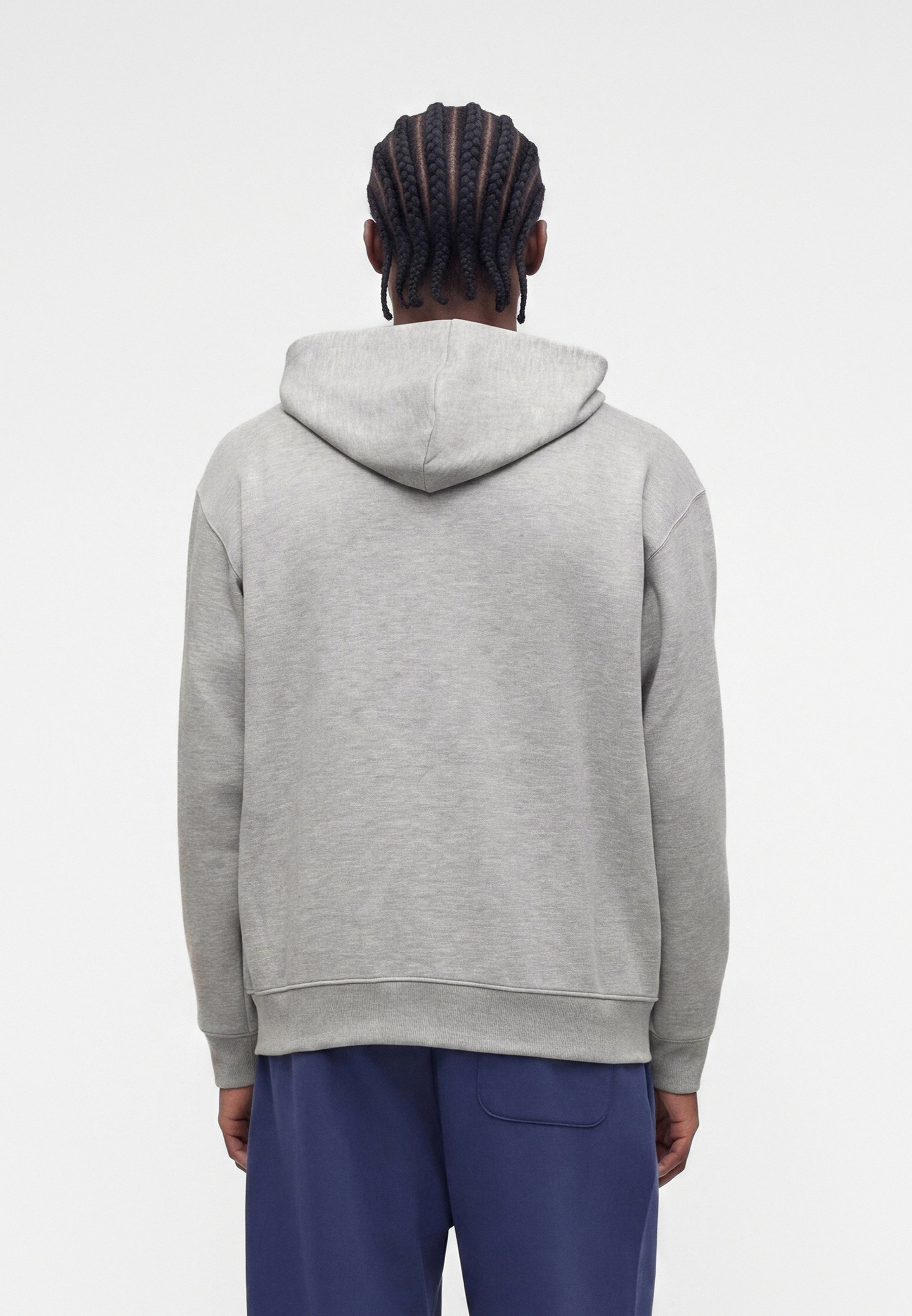 Persona con el pelo trenzado que lleva una sudadera gris con capucha y pantalones azules, de pie de espaldas sobre un fondo claro y liso.