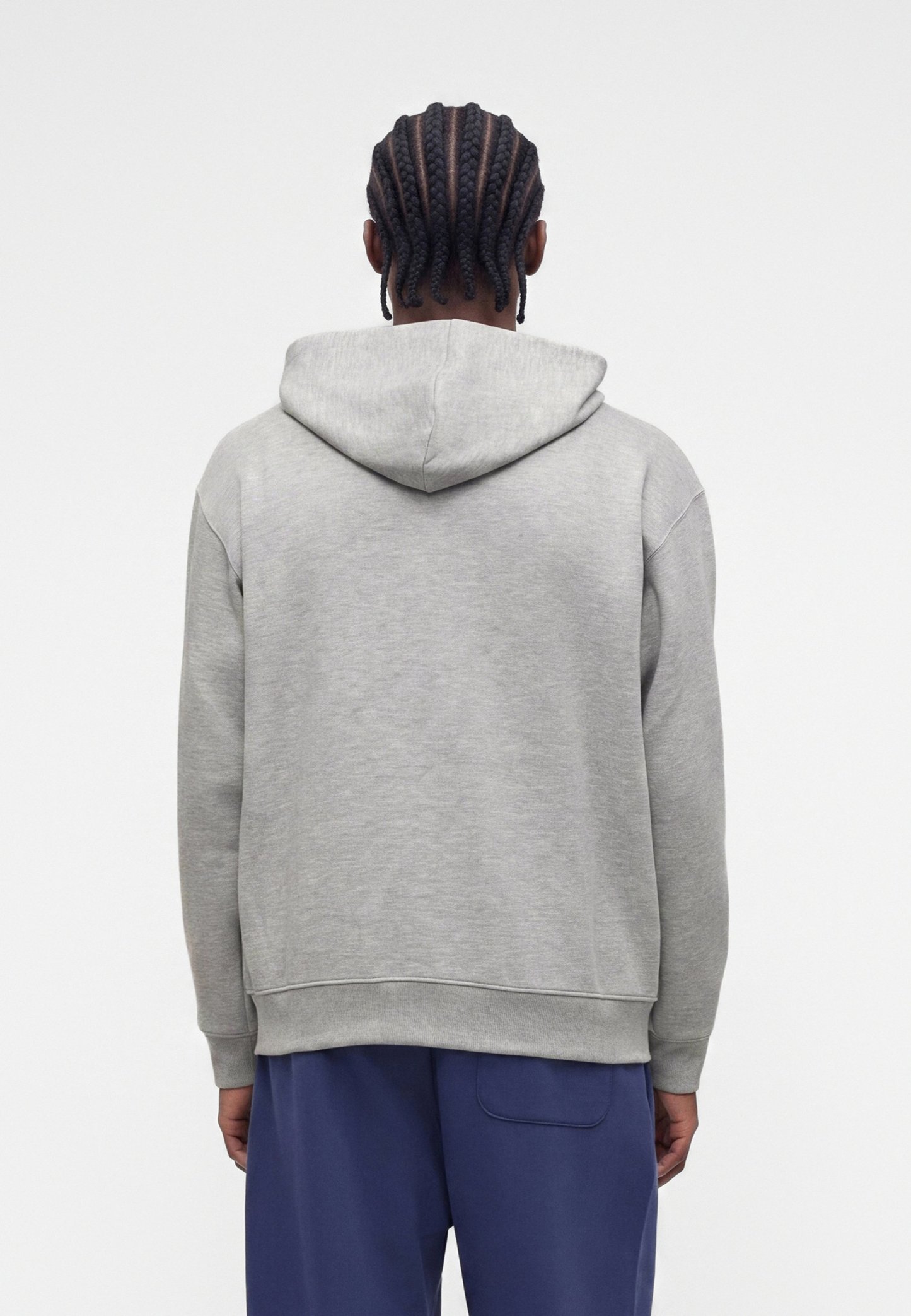 Persona con el pelo trenzado que lleva una sudadera gris con capucha y pantalones azules, de pie de espaldas sobre un fondo claro y liso.