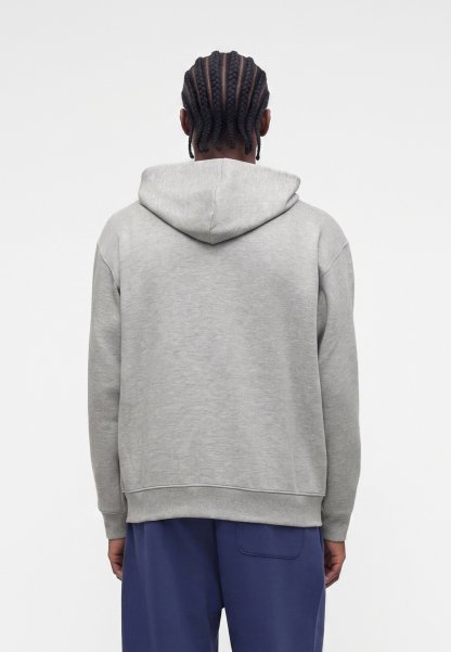 Persona con el pelo trenzado que lleva una sudadera gris con capucha y pantalones azules, de pie de espaldas sobre un fondo claro y liso.