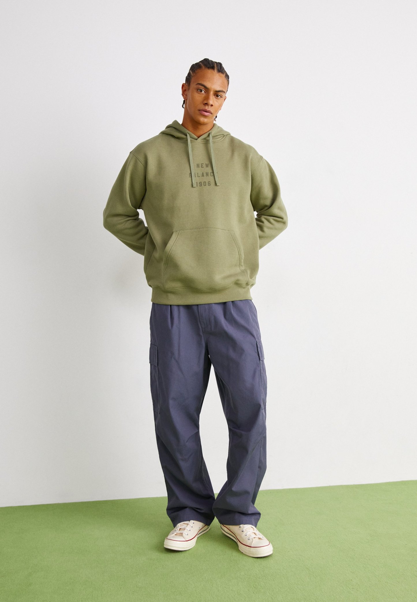Sudadera verde oliva con bolsillo frontal y capucha, combinada con pantalones cargo de corte holgado en color navy que cuentan con bolsillos laterales y zapatillas color crema.