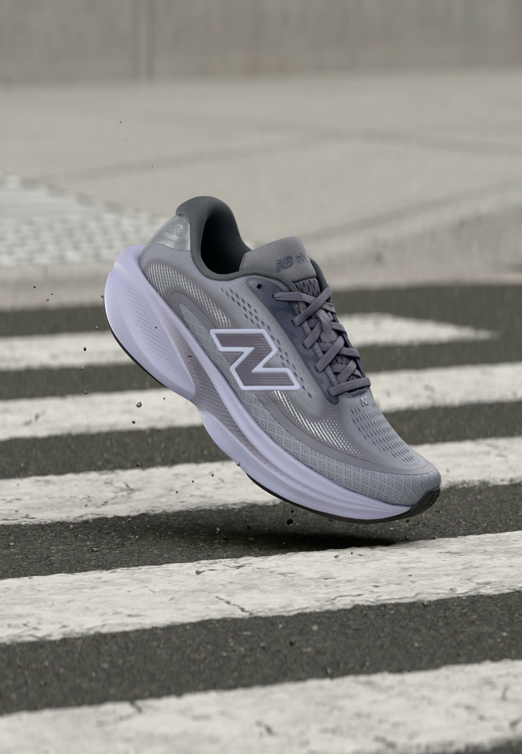 Zapatilla deportiva gris New Balance en el aire sobre un paso de peatones en asfalto, mostrando el lateral con un gran logo "N" y suela blanca acolchada.