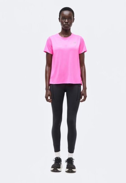 Mujer de pie mirando hacia adelante, vistiendo una camiseta de manga corta de color rosa brillante, mallas negras, calcetines blancos y zapatillas de correr negras.