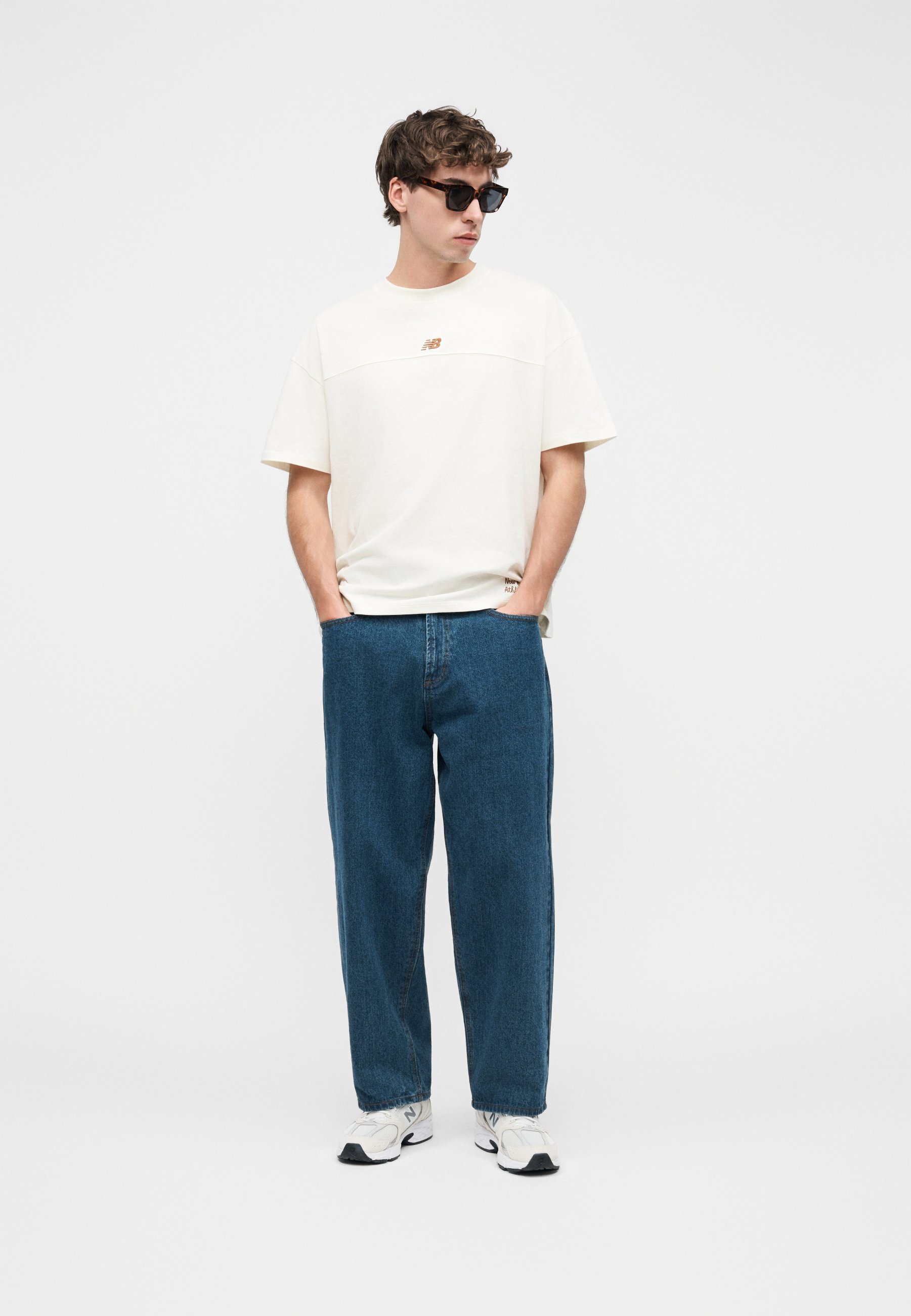 Hombre con camiseta blanca de gran tamaño, vaqueros azules holgados, zapatillas blancas y gafas de sol, de pie con las manos en los bolsillos sobre fondo blanco.