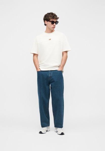 Hombre con camiseta blanca de gran tamaño, vaqueros azules holgados, zapatillas blancas y gafas de sol, de pie con las manos en los bolsillos sobre fondo blanco.