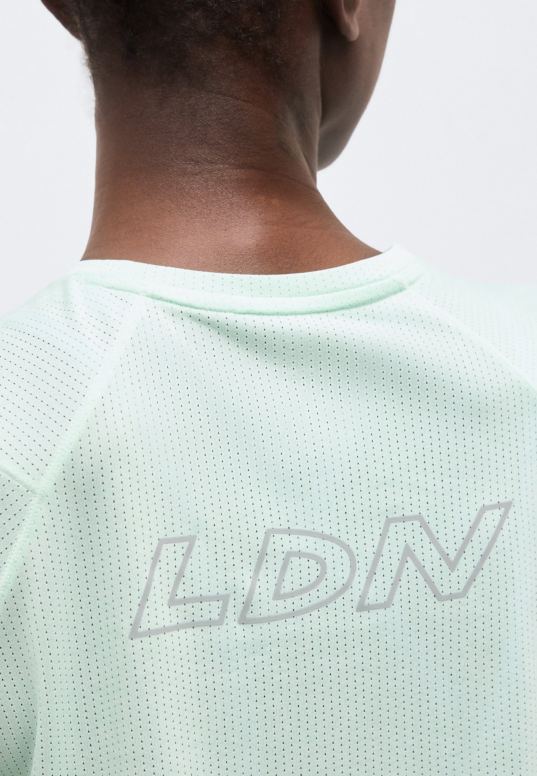 Primer plano del cuello y la parte superior de la espalda de una persona que lleva una camisa de tela perforada de color verde claro con las letras "LDN" impresas en letras reflectantes.