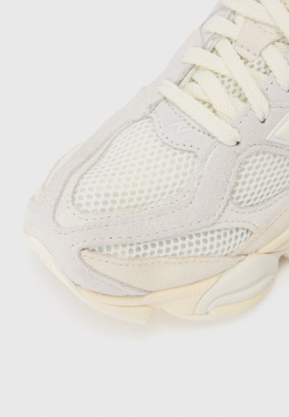 Zapatilla gris claro con paneles de malla y acentos de ante. Cuenta con una suela de goma texturizada y cordones beige, diseñada para comodidad y soporte.