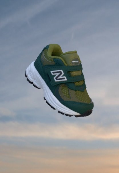Zapatilla New Balance verde y verde azulado oscuro con suela blanca, flotando contra un fondo de cielo nublado.