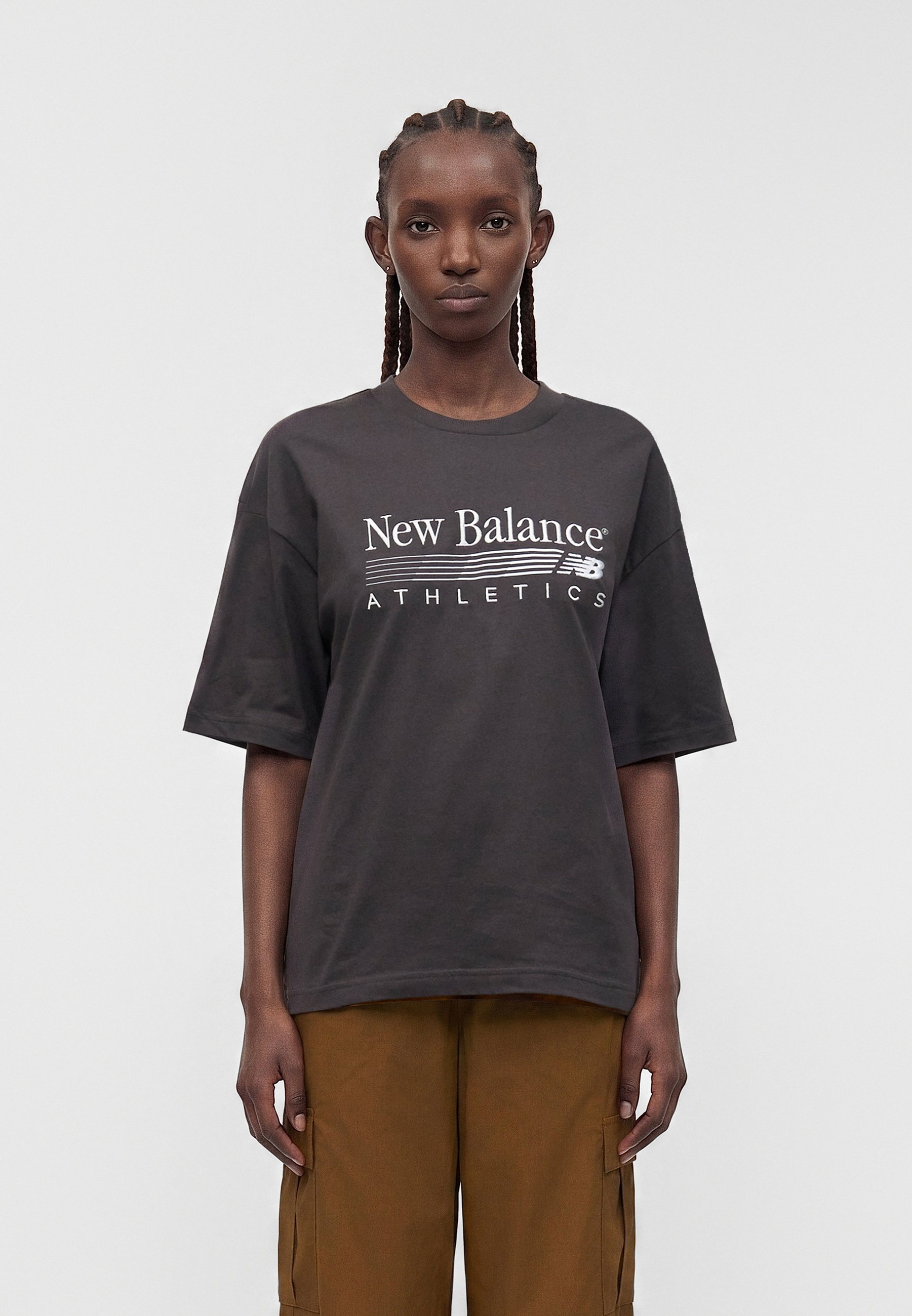 Mujer con el pelo trenzado que lleva una camiseta negra suelta de New Balance Athletics y pantalones cargo marrones, de pie frente a un fondo liso.