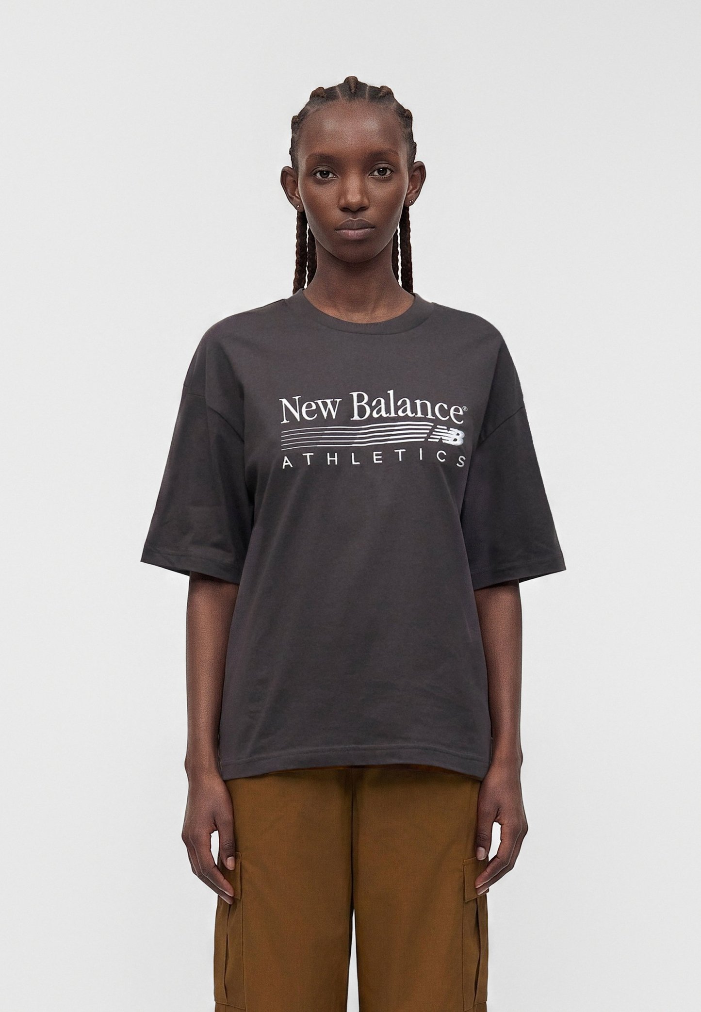 Mujer con el pelo trenzado que lleva una camiseta negra suelta de New Balance Athletics y pantalones cargo marrones, de pie frente a un fondo liso.