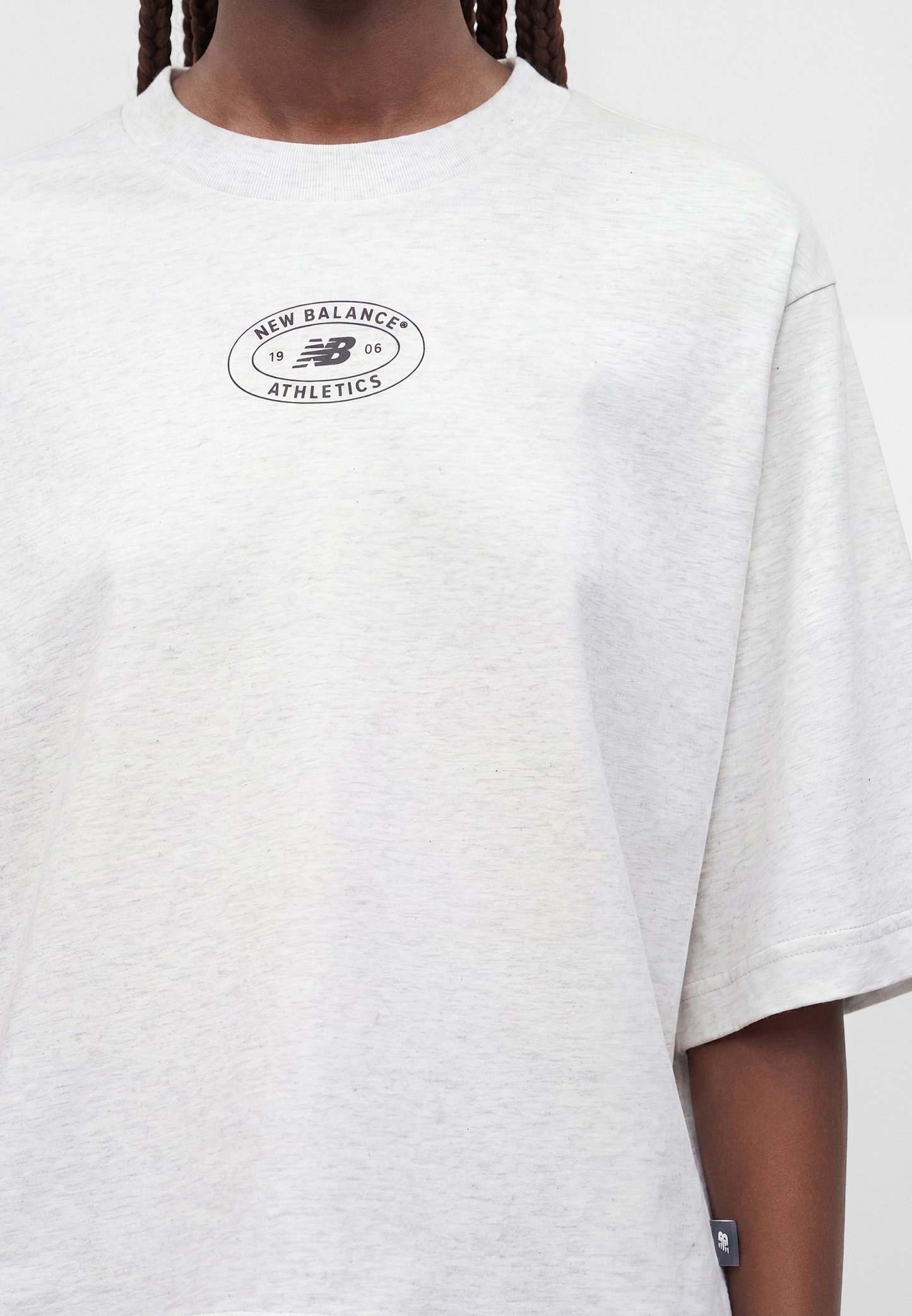 Primer plano de una persona que lleva una camiseta New Balance Athletics de color gris claro con un pequeño logo negro en el pecho y una etiqueta en el lateral.
